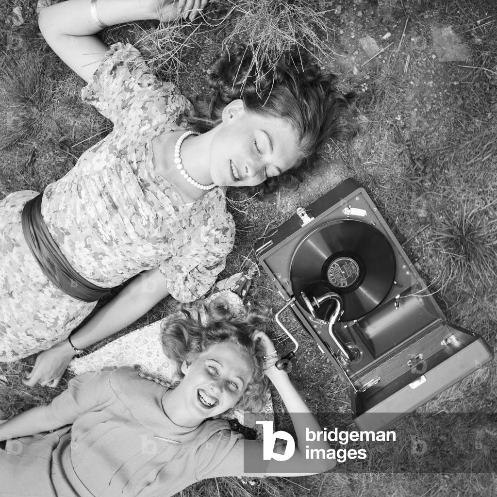 Two young women lying in a lawn and listening to the lastest hits from their Electrola gramophone, Germany 1930s (b/w photo)
