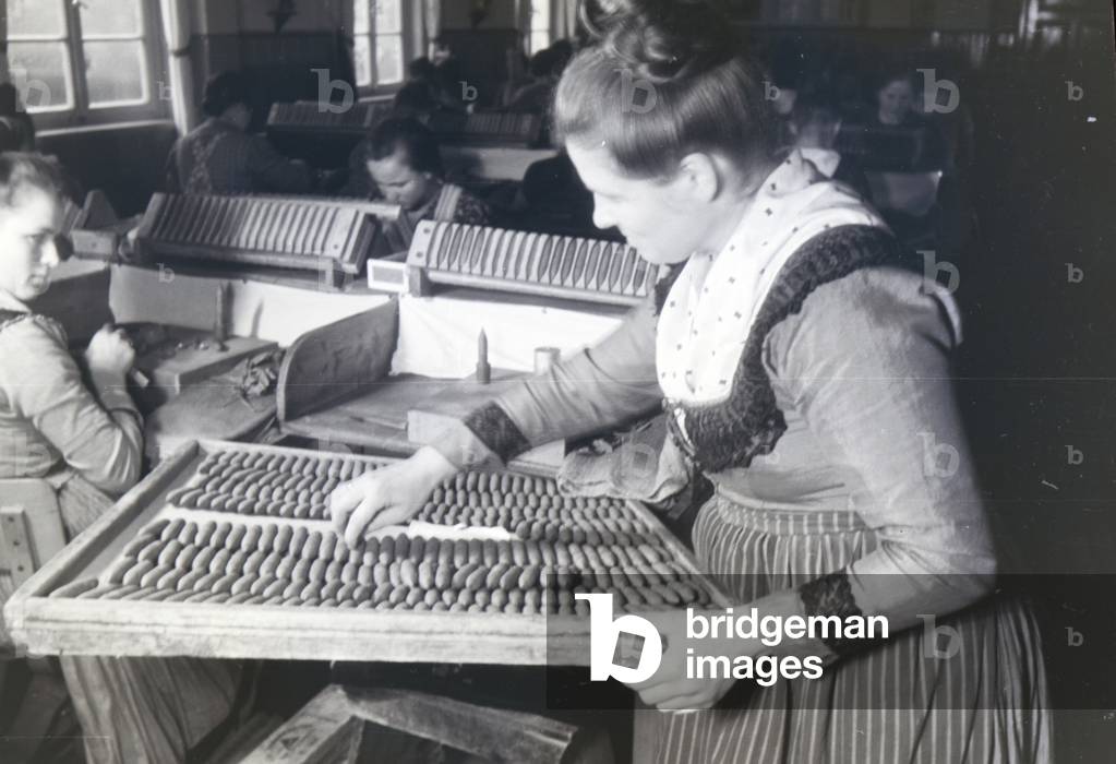A female cigar roller wearing a Marburg garb, costume is counting the finished hand-rolled cigars that are sorted on a big wooden plate, near Marburg, Germany 1930s (b/w photo)