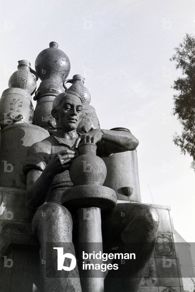 The statue of a master potter shaping a vase on the pottery wheel with his hands, with many decorated stoneware jars behind him, Kannenbäckerland, Germany 1930s (b/w photo)