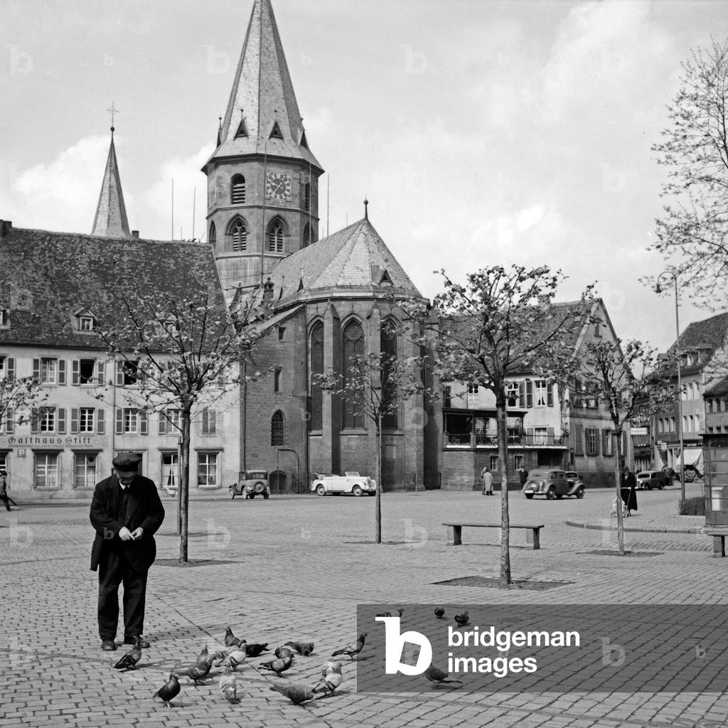 A man feeding pigions at the square in front of St Martin's and St Mary's collegiate church at Kaiserslautern, Germany 1930s (b/w photo)