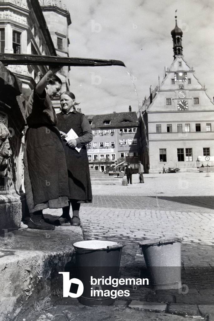 Two women having a chat while the bucktes are filling with water from the fountain placed on the market square near the town hall and Ratstrinkstube in Rothenburg ob der Tauber, Germany 1930s (b/w photo)