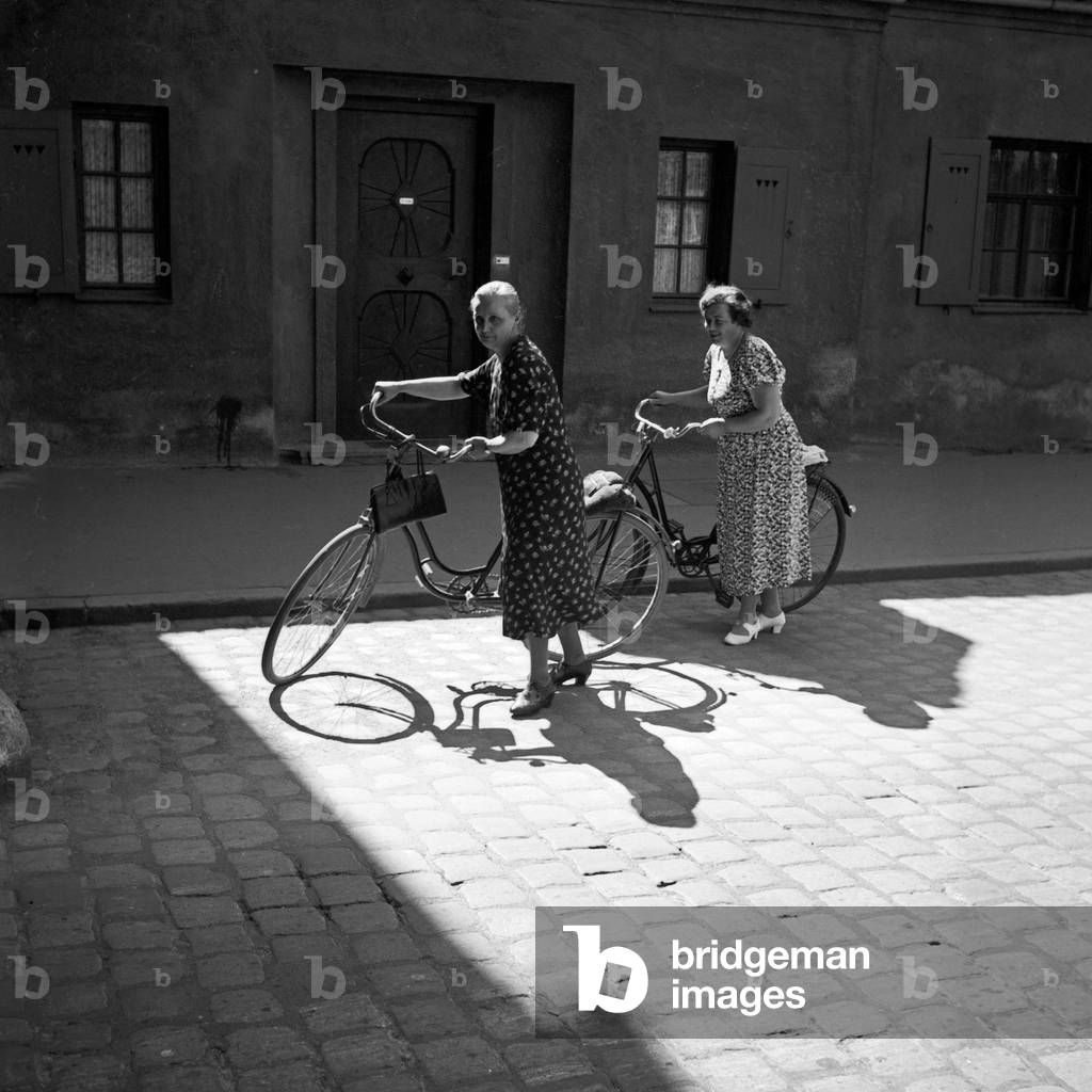 Two ladies pushing their bicycles through the old city of Augsburg, Germany 1930s (b/w photo)