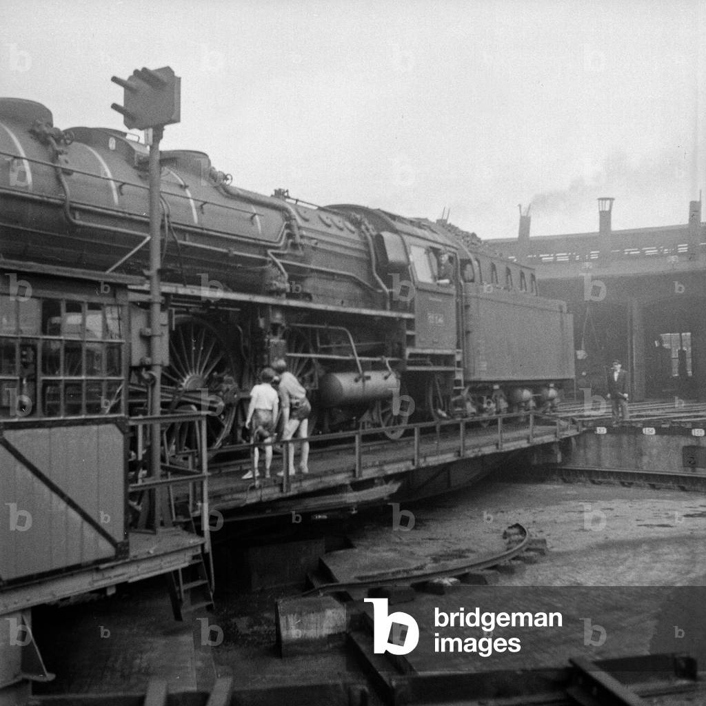 Two boys gazing at a steam engine of type 03 on a pallet in front of an engine shed at Hamburg depot, Germany 1950s