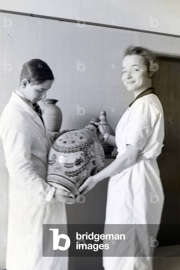 Two students of the College for Ceramics in Höhr-Grenzhausen presenting the ceramics exhibited in the hallway of the school, Germany 1930s (b/w photo)