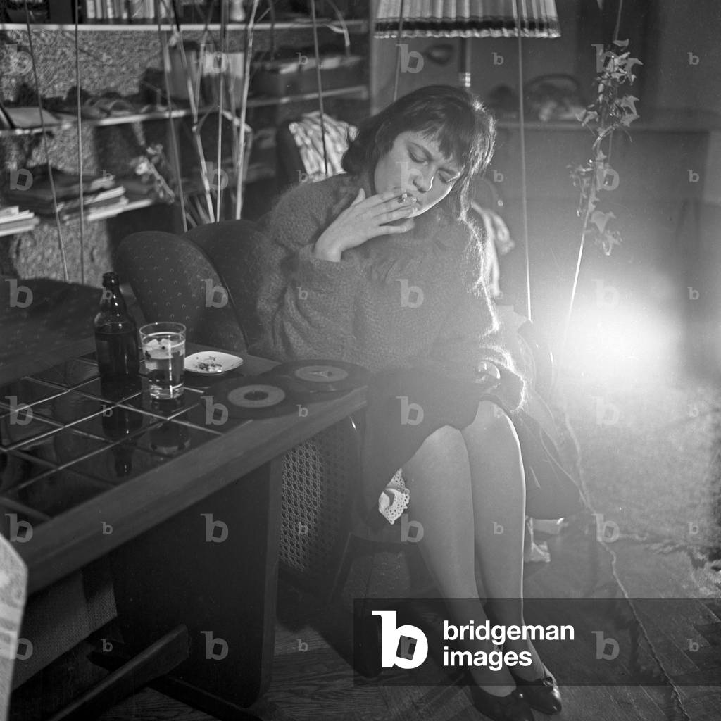 A young woman relaxing with a glass of beer while listening to some record discs at the living room, Germany 1950s