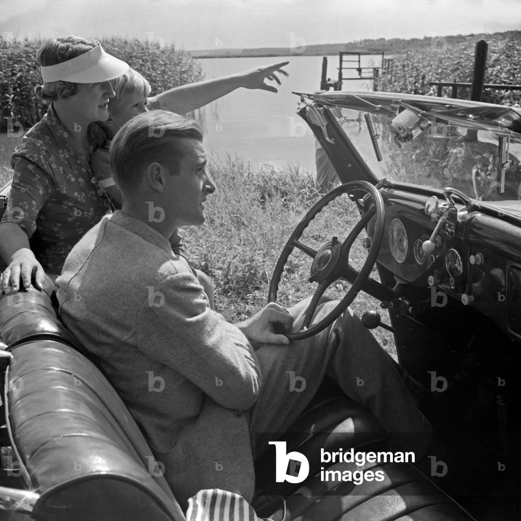 A man sitting on the steering wheel of his convertible while his family has a look at a lake, Germany 1930s (b/w photo)