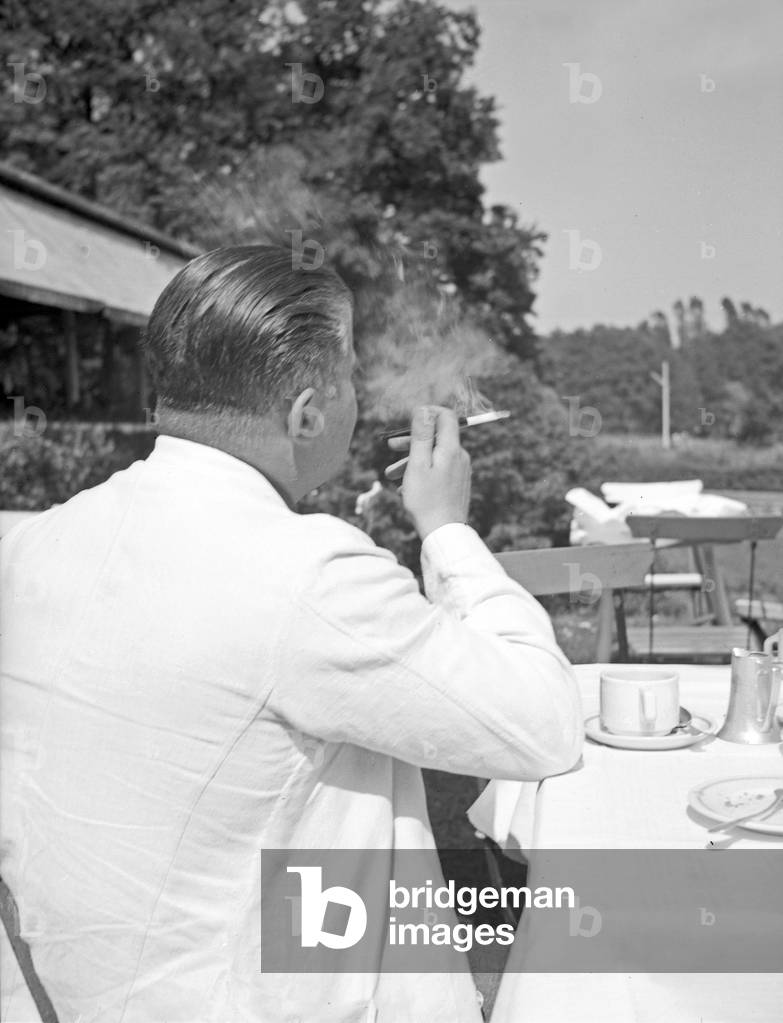 A man sitting in the open air gastronomy of a restaurant smoking a cigarette, Germany 1930s (b/w photo)