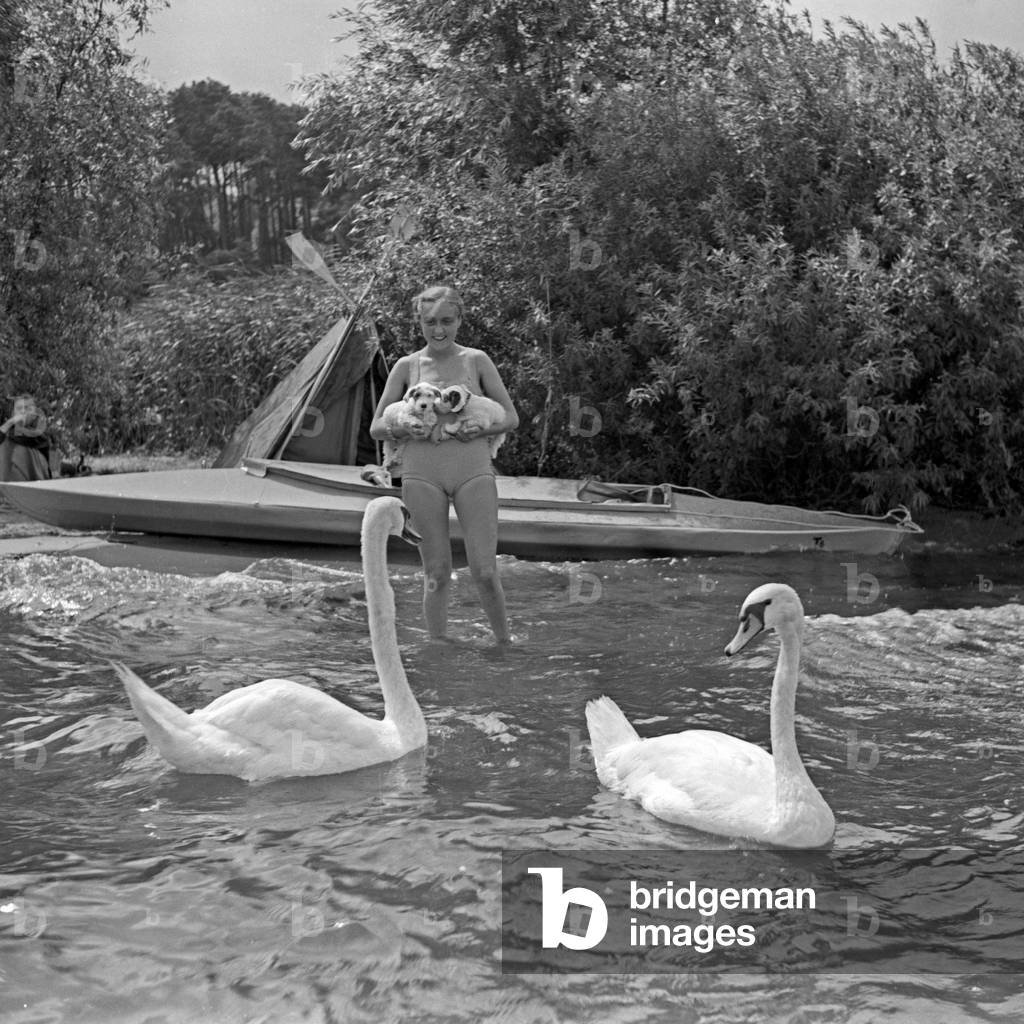 Advertising für a Klepper foldboat and tent: a young woman playing with two puppies in front of a boat and a tent at the shore of a lake, Germany 1930s (b/w photo)