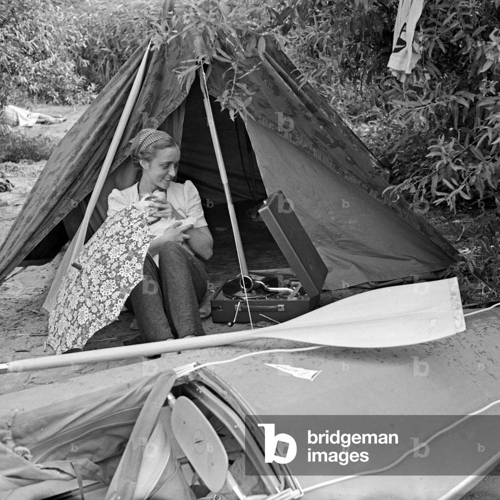 Advertising für a Klepper foldboat and tent: a young woman sitting with a puppy in front of a boat and a tent at the shore of a lake, Germany 1930s (b/w photo)