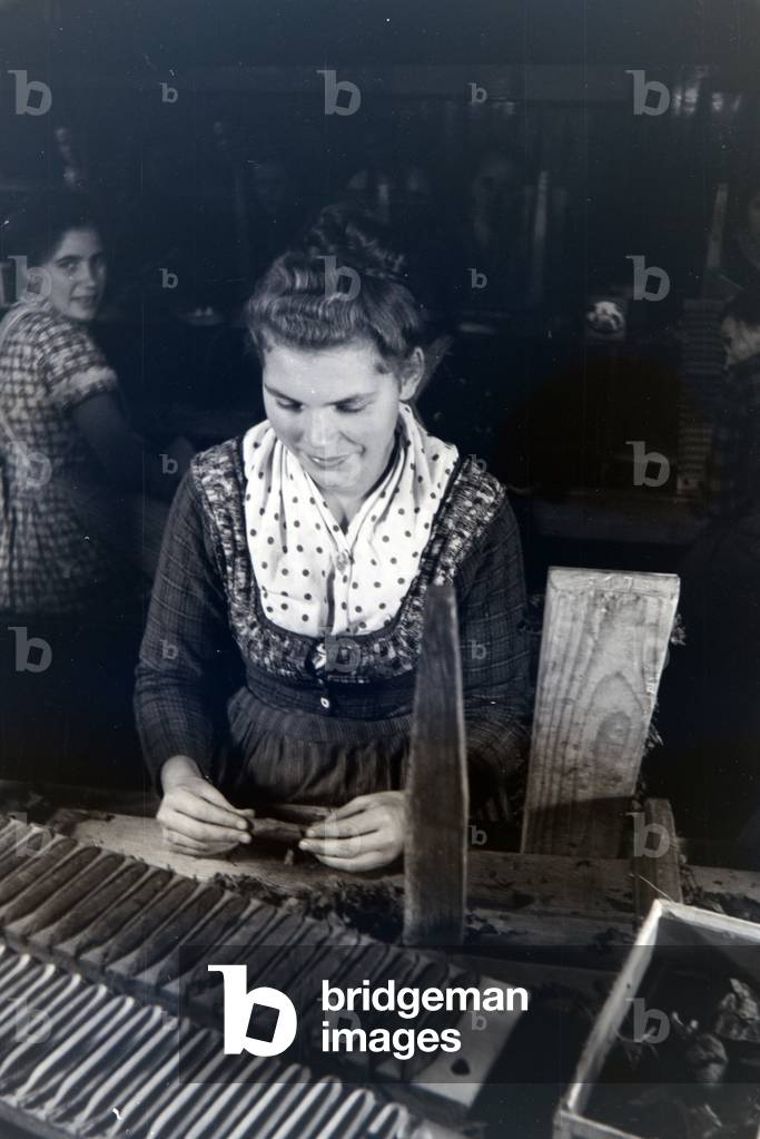 A female cigar roller wearing a Marburg garb, costume at work in a cigar fabric near Marburg, Germany 1930s (b/w photo)
