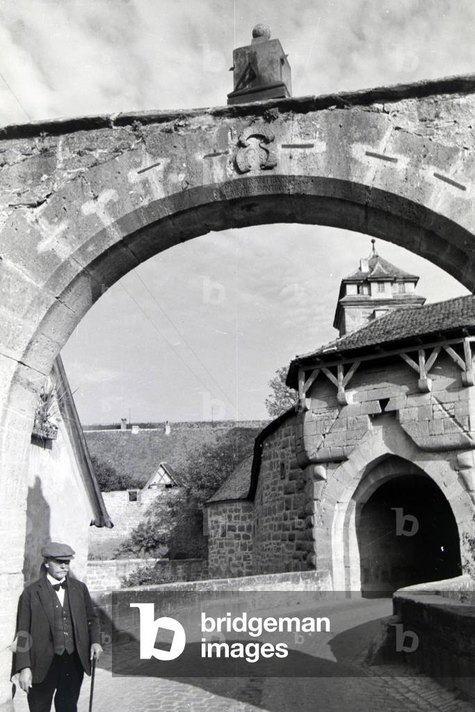 An elderly man with a walking stick posing under an archway in Rothenburg ob der Tauber, Germany 1930s (b/w photo)