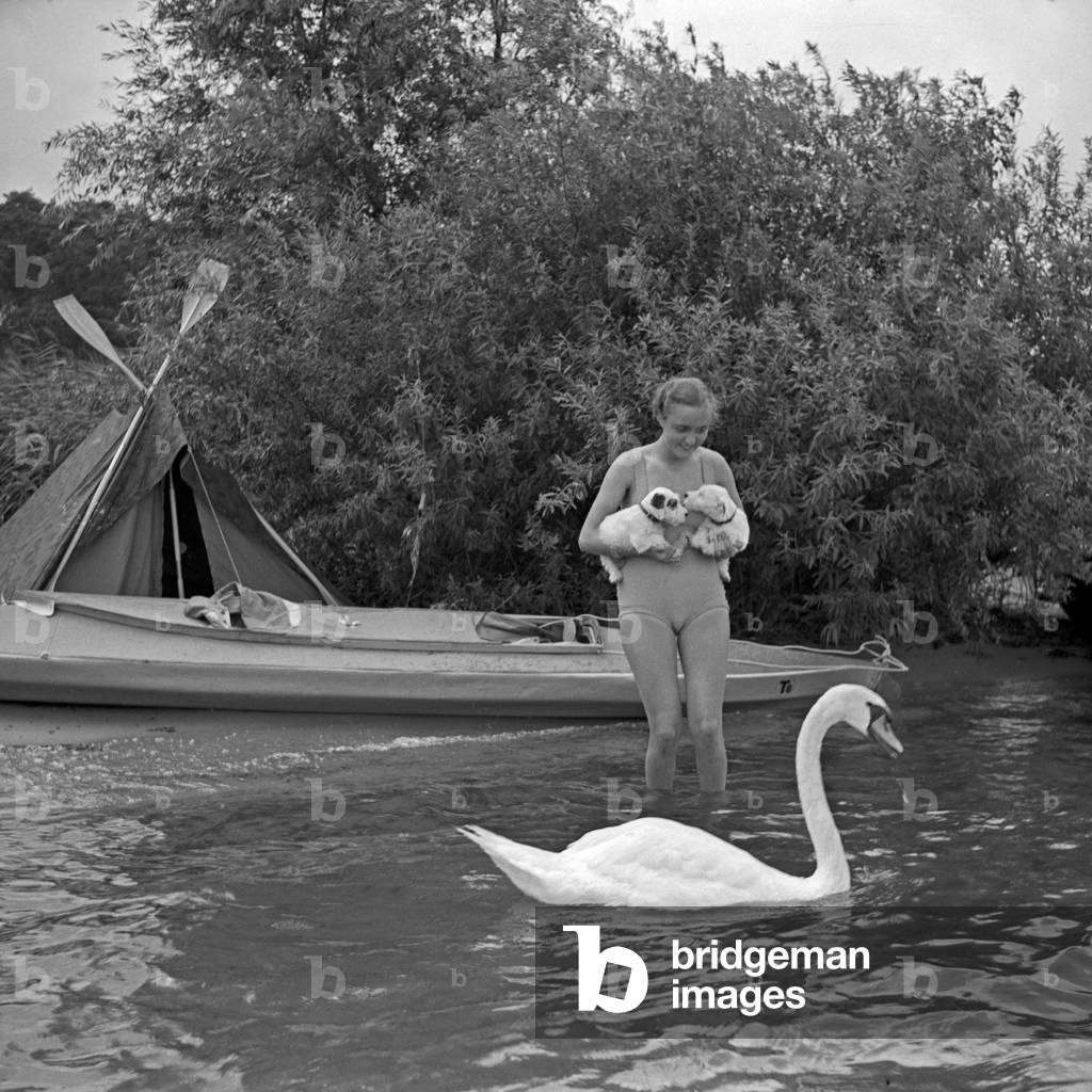 Advertising für a Klepper foldboat and tent: a young woman playing with two puppies in front of a boat and a tent at the shore of a lake, Germany 1930s (b/w photo)