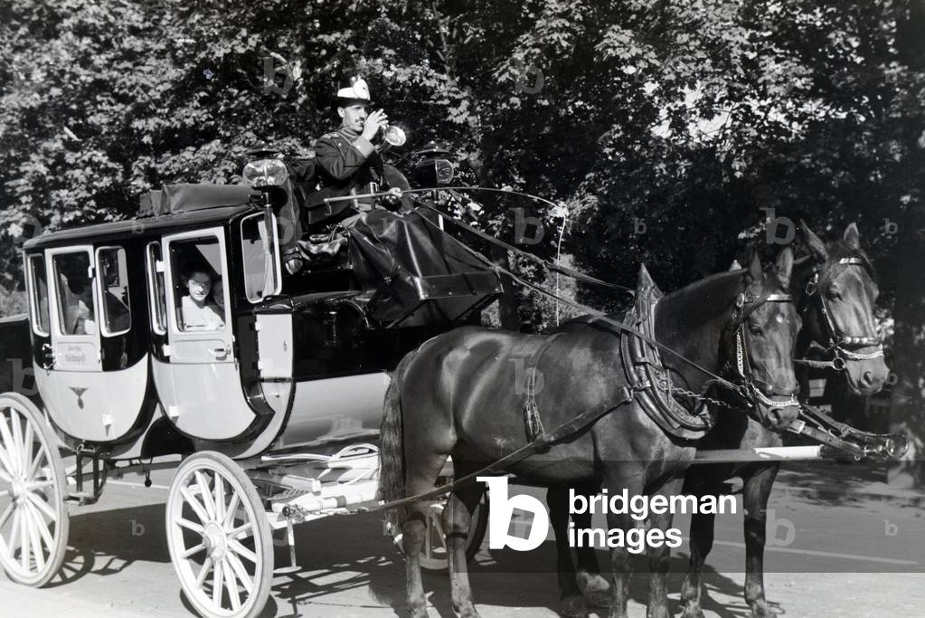 A coachman wearing a traditional uniform trumpeting from his coachman´s seat while his passengers are peeking out the window curiously, Bad Blankenburg, Germany 1930s (b/w photo)