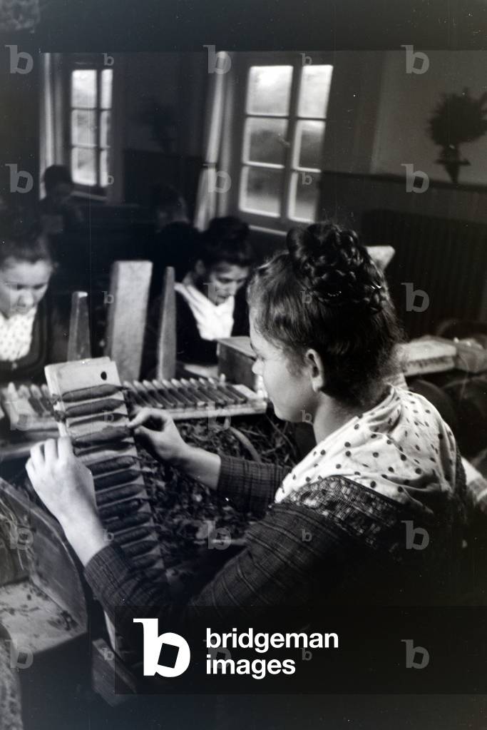 A female cigar roller wearing a Marburg garb, costume at work in a cigar fabric near Marburg, Germany 1930s (b/w photo)