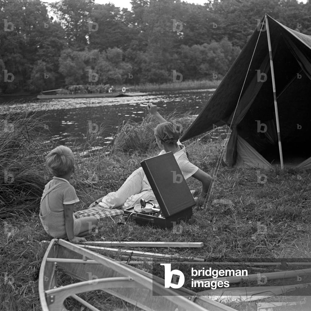 A young woman and a boy sitting in front of a tent and a folding boat of the brand Klepper, listening to music played by an Electrola Koffer 106 suitcase gramophone, Germany 1930s (b/w photo)