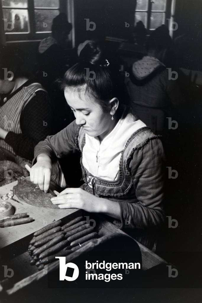 A female cigar roller wearing a Marburg garb, costume at work in a cigar fabric near Marburg, Germany 1930s (b/w photo)
