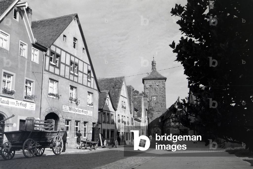 A paving stone alley leading toward the Sieberstor, Siebers Gate in Rothenburg ob der Tauber, past old carriages and half-timbered houses, Germany 1930s (b/w photo)