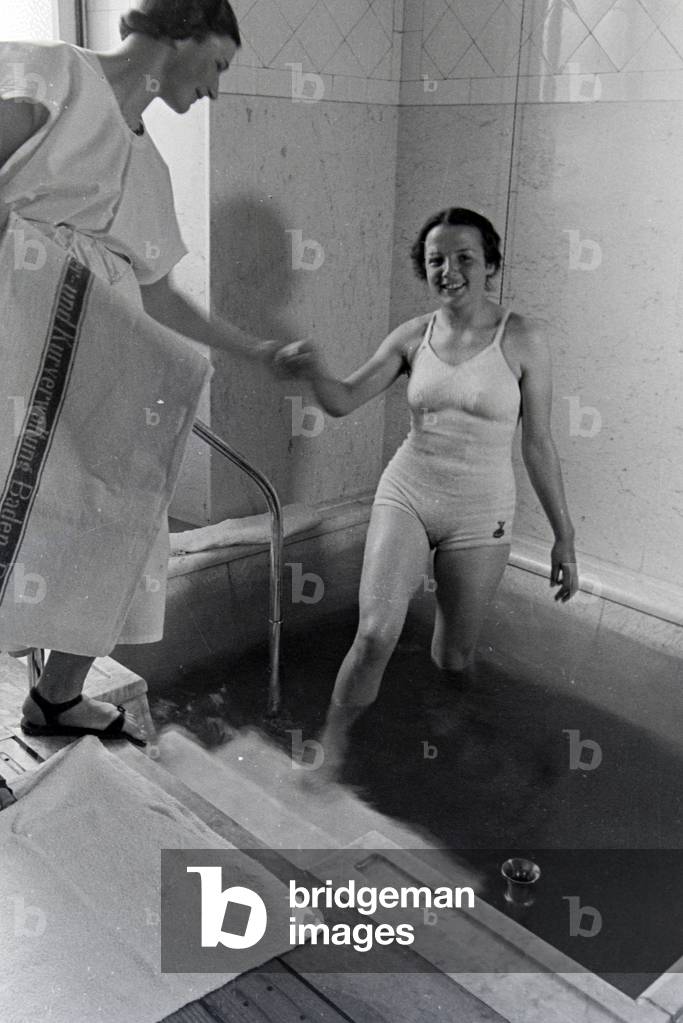 An employee of the Caracalla Therme in Baden-Baden helps a young woman getting out of the hot water pool, Germany 1930s (b/w photo)