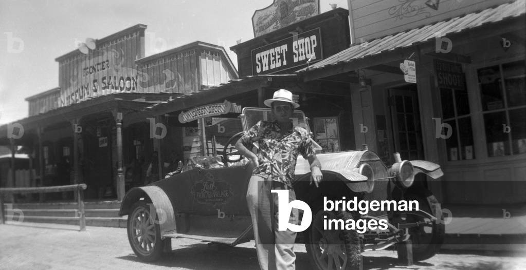 A man standing in front of his vintage car on the street of a Western town, 1954