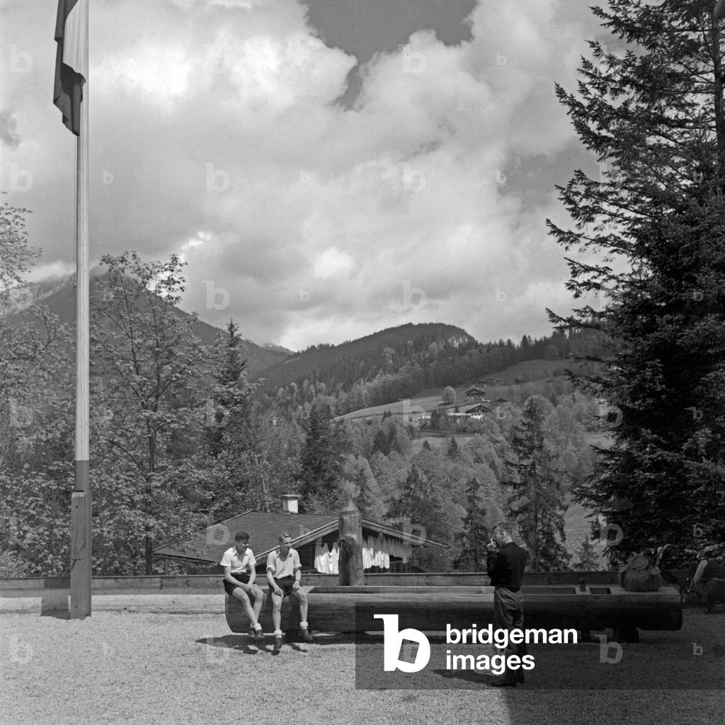 A boy taking a picture of two other boys in front of the mountain range at the courtyard of the Adolf Hitler youth hostel at Berchtesgaden, Germany 1930s