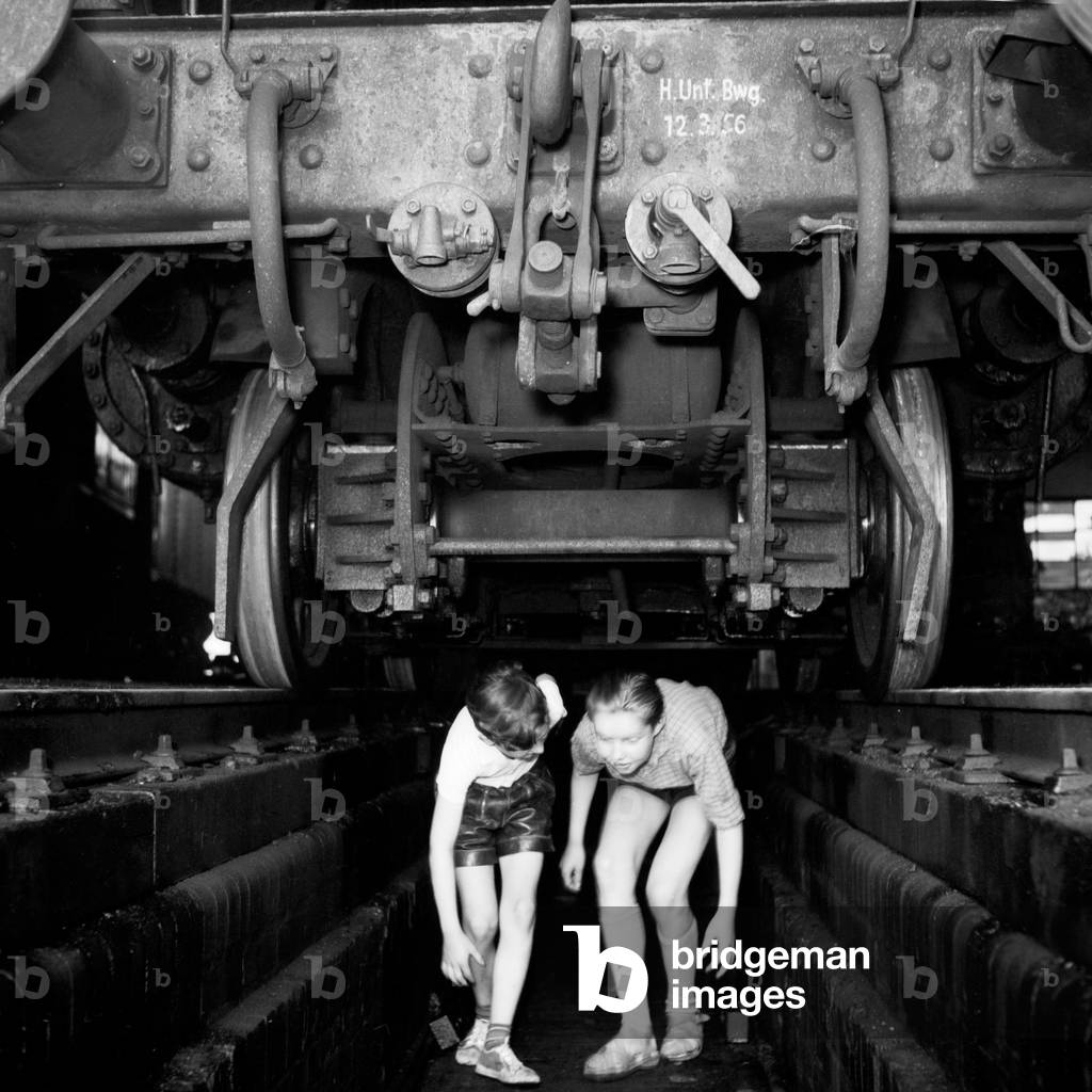 Two boys at a slag pit under a steam engine of type 03 in an engine shed at Hamburg depot, Germany 1950s