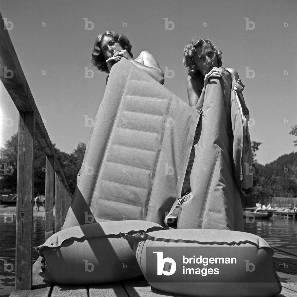 Bathing guests blowing upi their air matresses at the shore of a lake in Austria, 1930s (b/w photo)