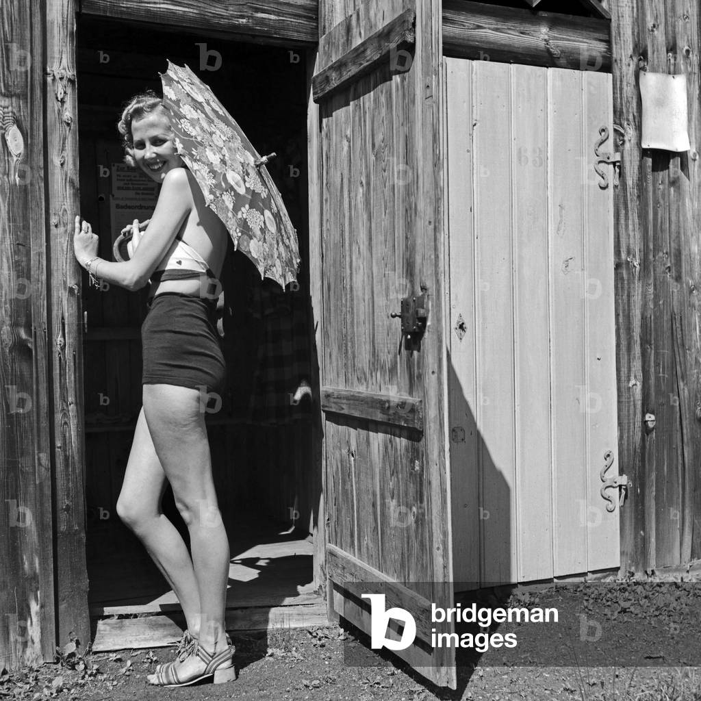 A young woman searing swimsuit with an umbrella at the entrance to a wodden hut, Germany 1930s (b/w photo)