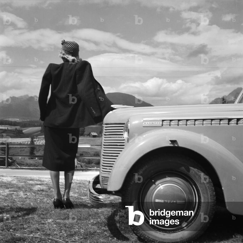 A young woman standing by the bonnet of an Opel model Olympia, Austria 1930s (b/w photo)