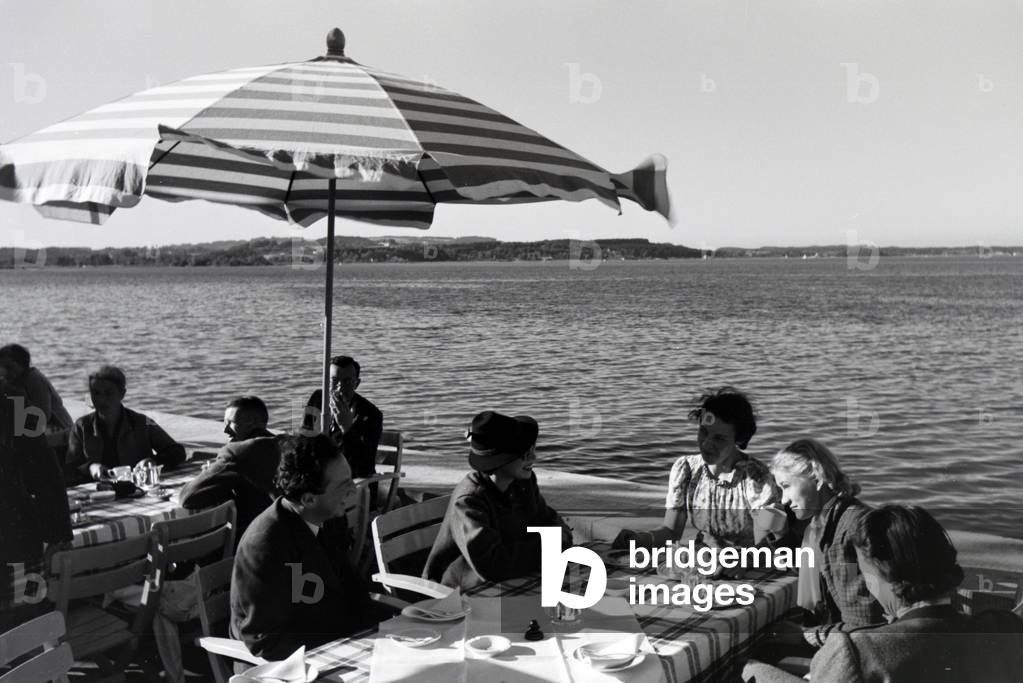 Guests of a café at the Chiemsee sitting under sun shades on the terrace, Germany 1930s (b/w photo)