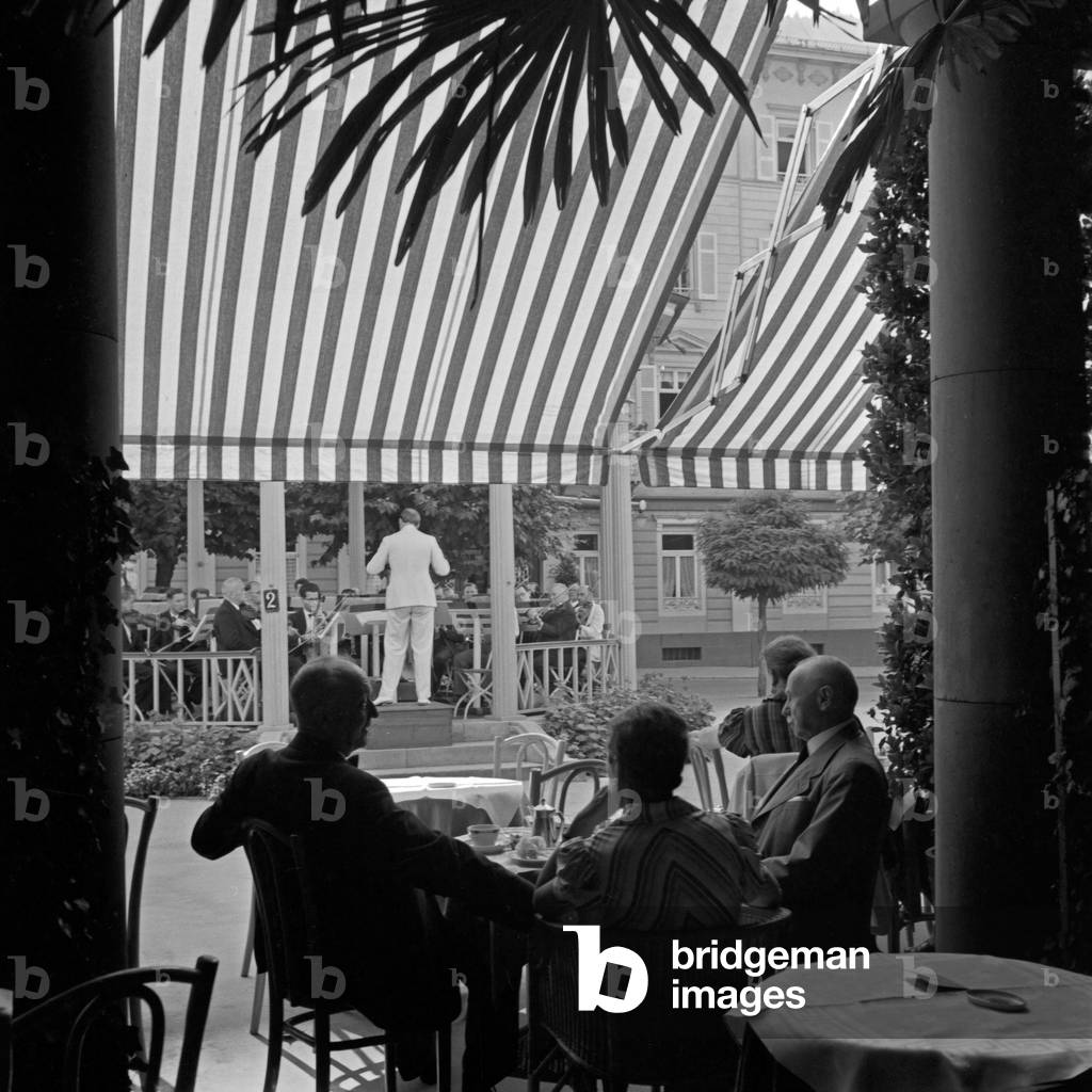 Spa guests sitting under a blind of an outdoor gastronomy and listening to the sounds of the spa orchestra at Wildbad in Black Forest, Germany 1930s (b/w photo)