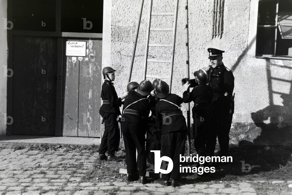 A group of junior firefighters is preparing a ladder under the guidance of a firefighter during a firefighter training, Germany 1930s (b/w photo)