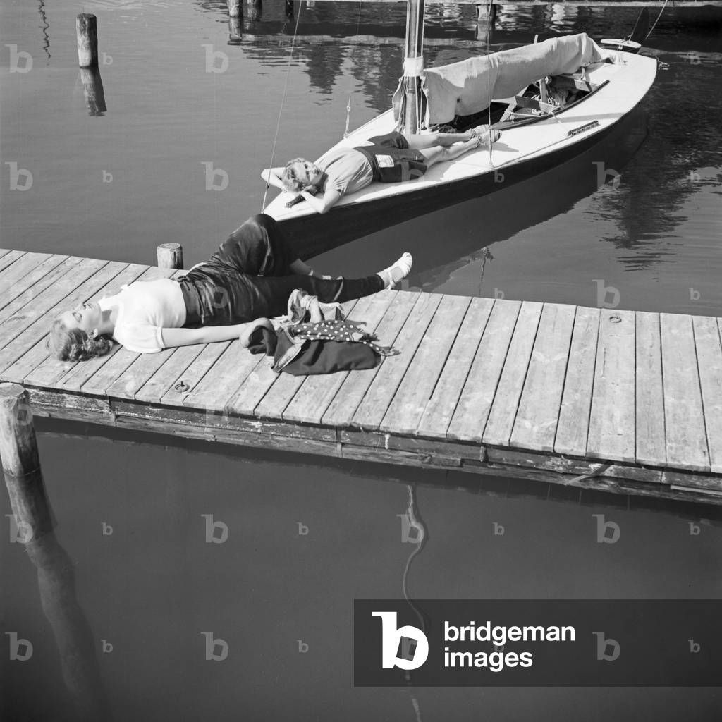 Two young woman tanning on the shore of a lake, one on a boardwalk, the other on a sailing boat, Germany 1930s (b/w photo)