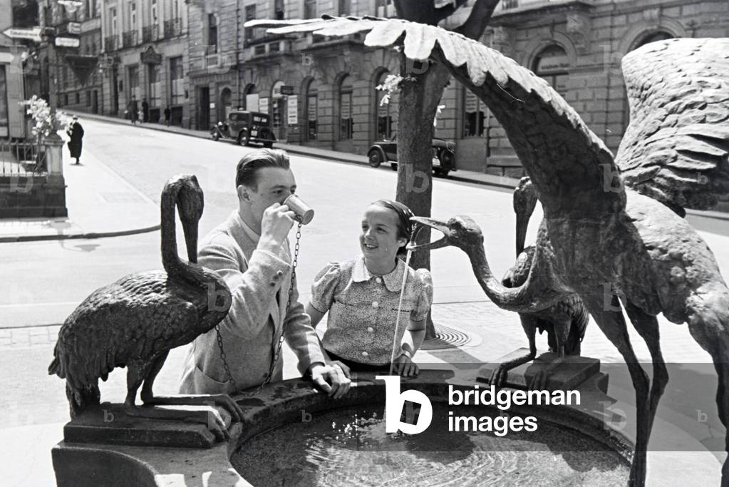 A young couple tastes the warm thermal water from the Reiherbrunnen in the Sophienstraße in the city centre of Baden-Baden, Germany 1930s (b/w photo)