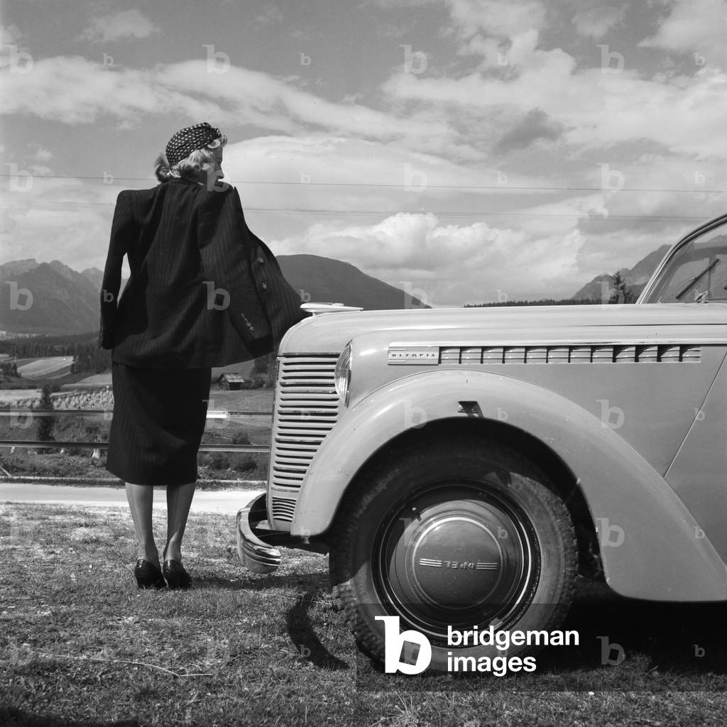 A young woman standing by the bonnet of an Opel model Olympia, Austria 1930s (b/w photo)