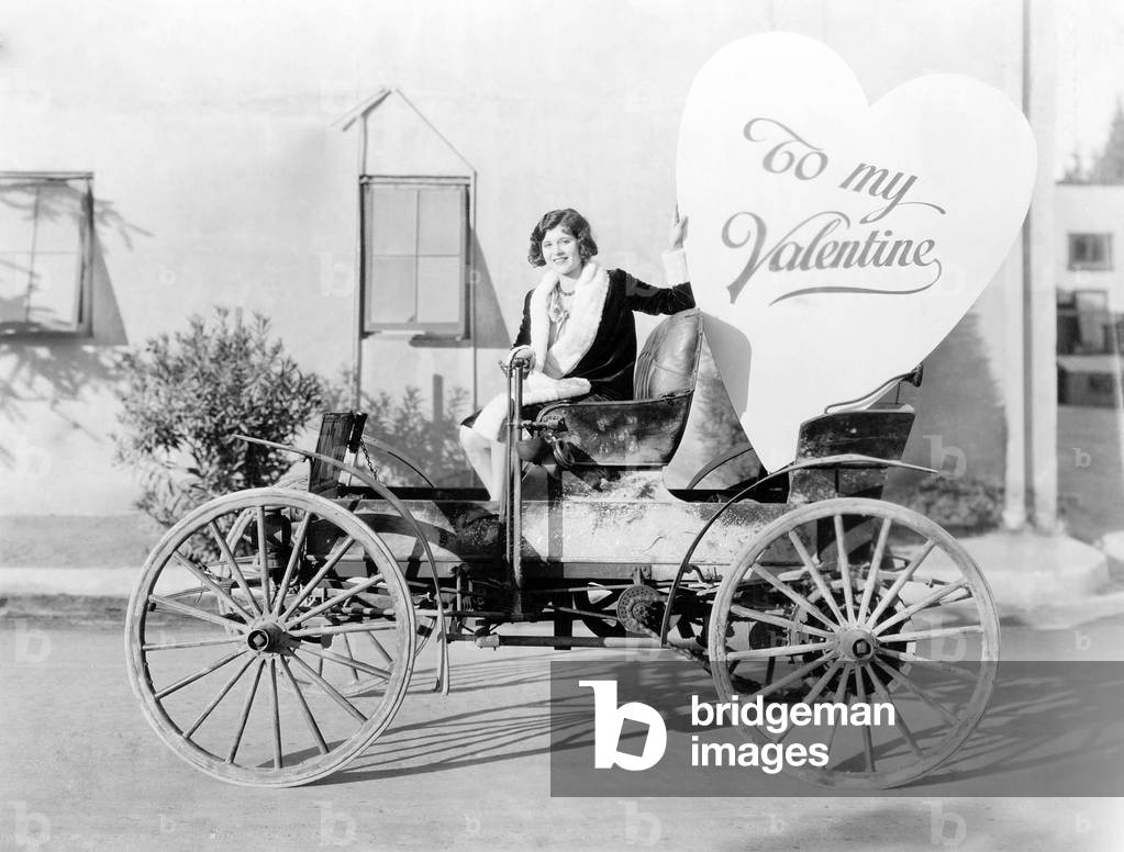 Young Woman Sitting on a Car Holding a Big Heart Shaped Sign