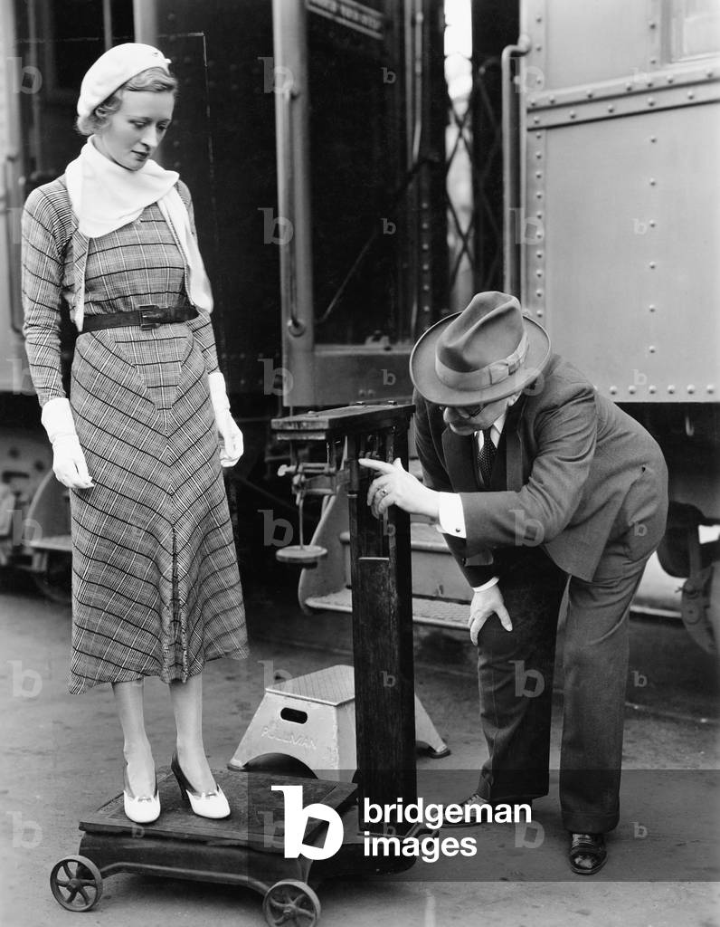 Profile of a Man Measuring Weight of a Woman Standing on a Weighing Scale in Front of a Train