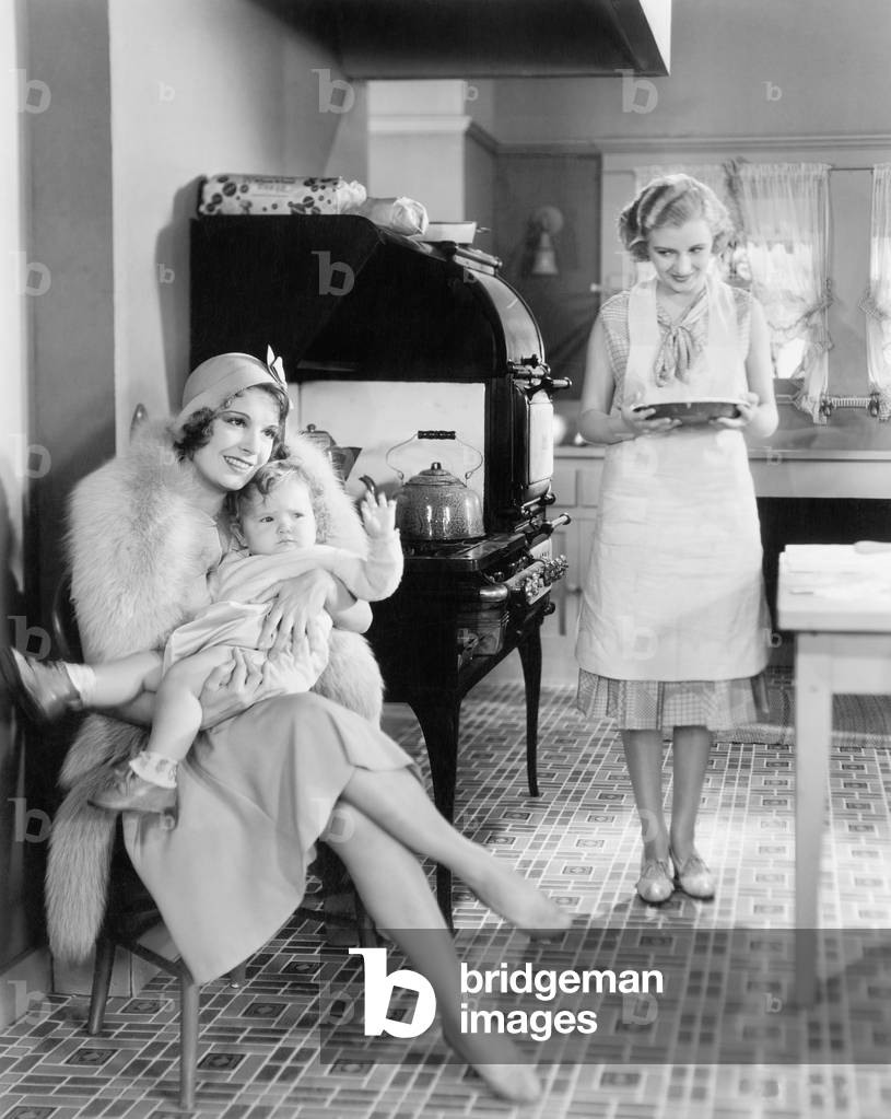 Elegant Woman Sitting with her Baby in a Kitchen While a Friend is Bringing a Pie
