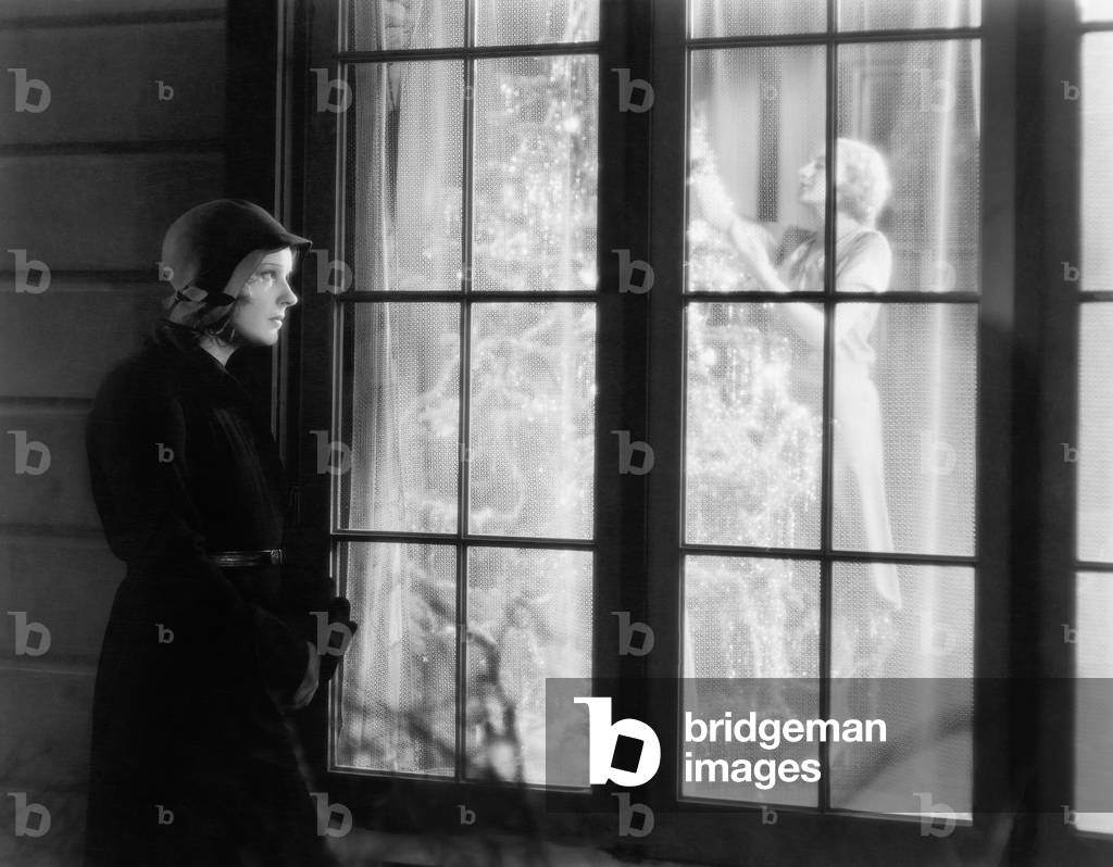 Woman Standing Outside of a Window Watching a Woman Trimming a Christmas Tree