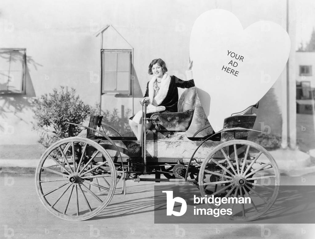 Young Woman Sitting on a Car Holding a Big Heart Shaped Sign