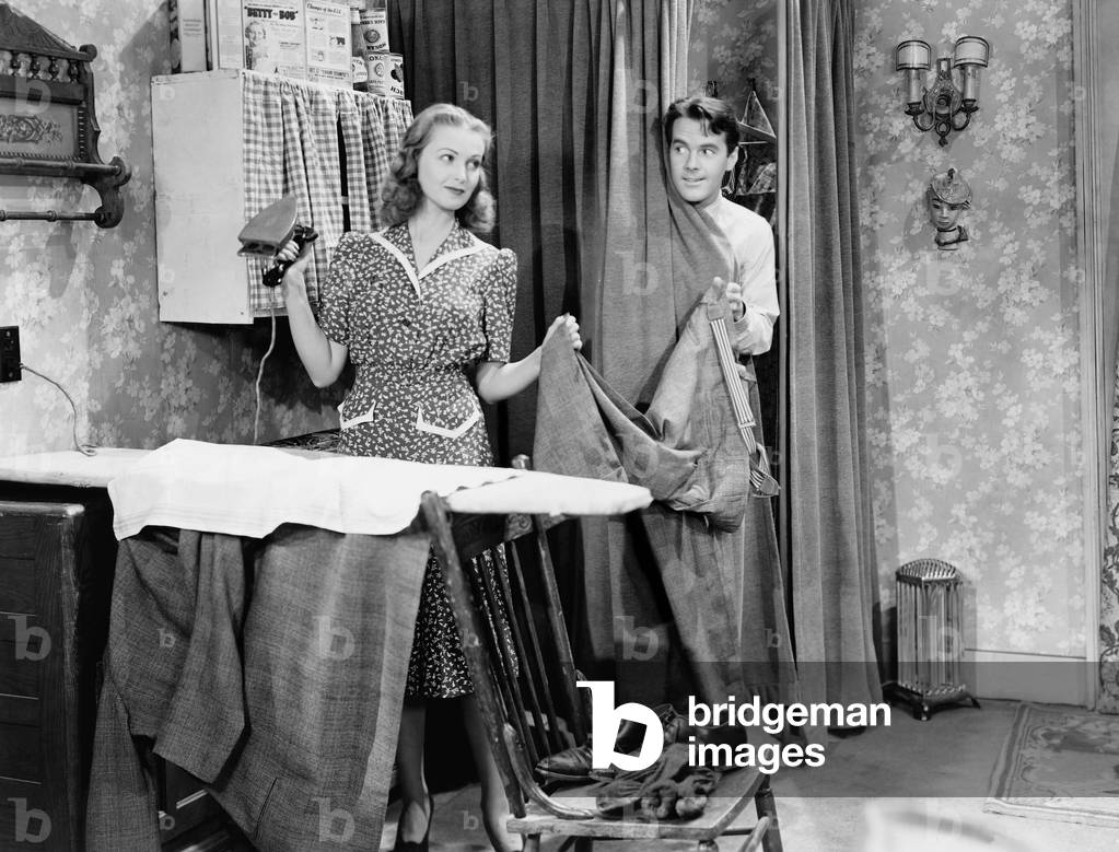 Man and Woman Standing in a Kitchen While She is Ironing his Pants and He is Behind a Curtain
