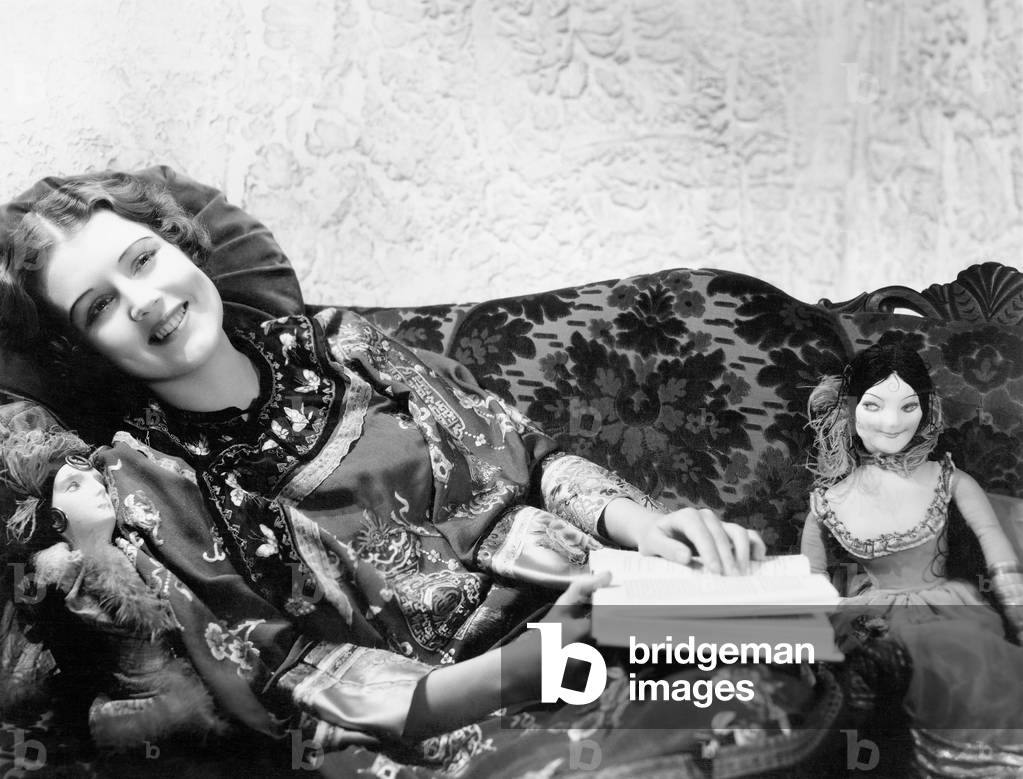 Woman in a Chinese Print Dress Lying on the Sofa with her Dolls and a Book