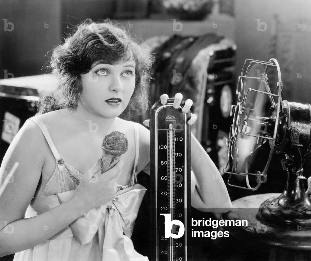 Young Woman Sitting Next to a Fan and a Thermometer Looking Hot and Eating an Ice Cream