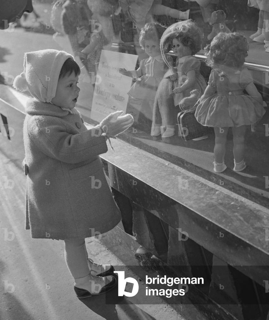 Young girl looking at dolls in the shop window of a department store in Paris, before Christmas, November 13, 1960 (b/w photo)