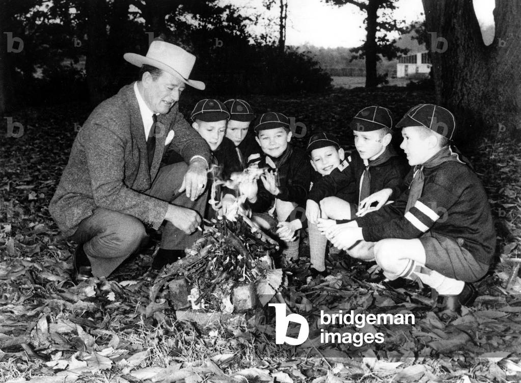 John Wayne showing how to light a fire outdoors to young scouts, 1960 (photo)