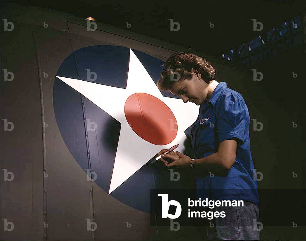 A candid view of one of the women workers touching up the U.S. Army Air Forces insignia on the side of the fuselage of a 