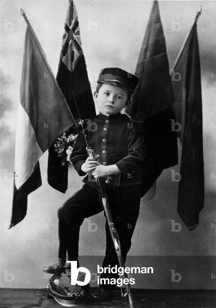 German defeat, 1918 : a boy, probably a on of a French soldier, posing with uniform, bayonet, the feet on a German helmet, end of ww1