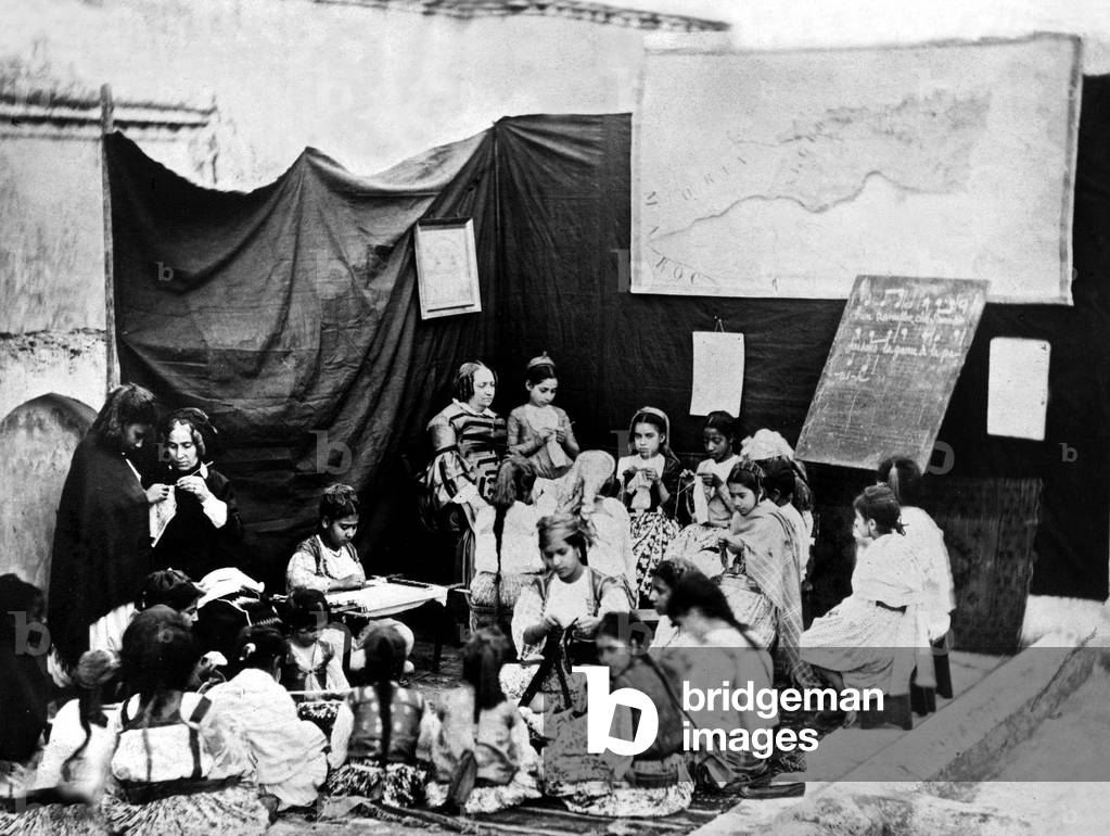 Teaching by european women to native little girls : they are learning sewing, knitting, we can see a map of north Africa, c. 1860