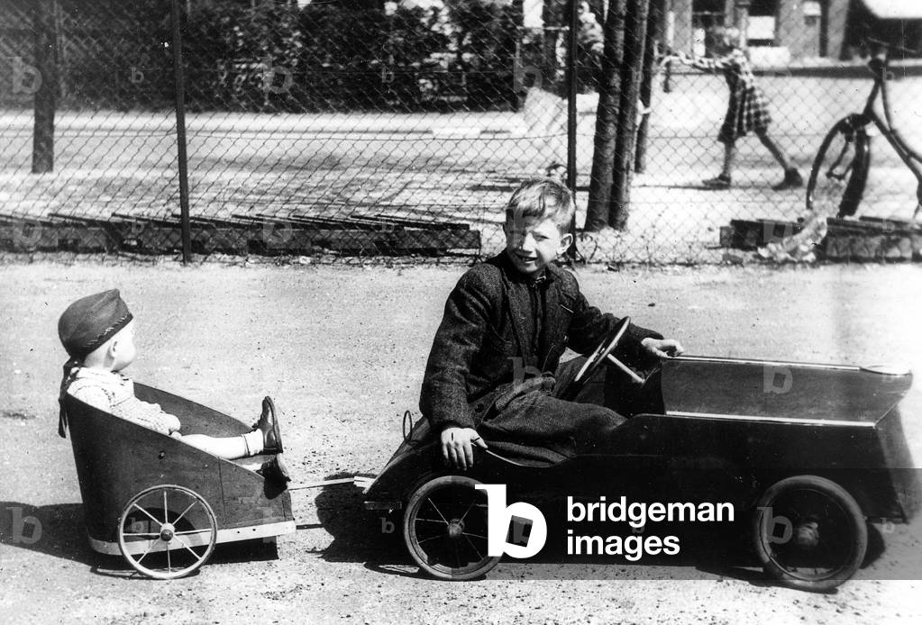 A boy in a small pedal car pulling his little sister in a trolley tied to car 40's