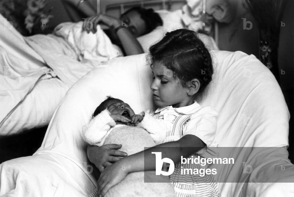 Little girl holding in her arms her sister while her mother has just given birth, 1949