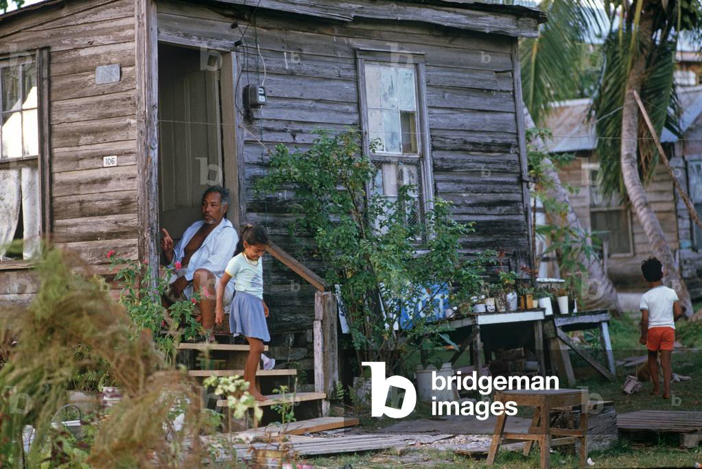 Belize uomo seduto nella porta di casa rivestita di legno con i bambini che giocano fuori, Belize City, Belize